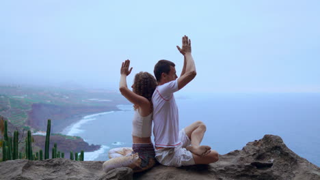 Amidst-mountain-views-and-the-ocean's-horizon,-a-man-and-woman-sit-on-a-rock-back-to-back,-immersed-in-meditation-and-yoga
